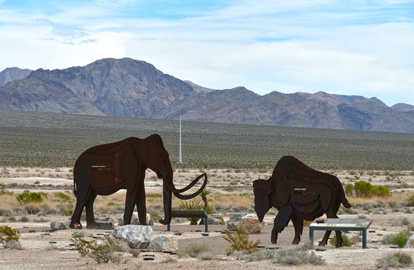 Metal sculptures of an elephant and a bison in a desert landscape with mountains in the background.