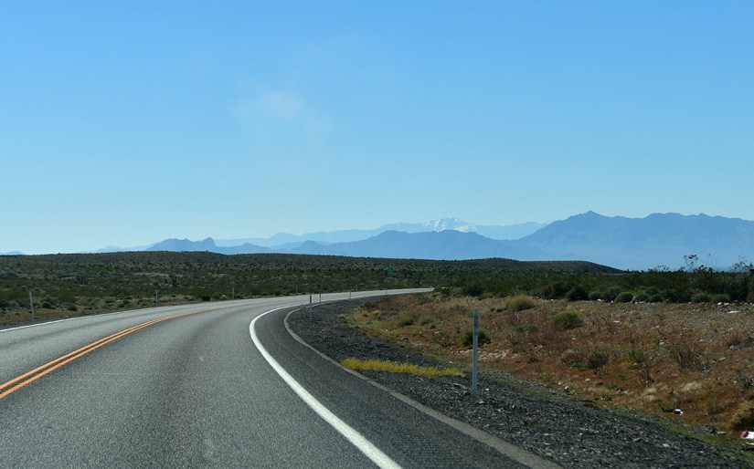 A winding road stretches through a desert landscape with mountains in the background under a clear blue sky.
