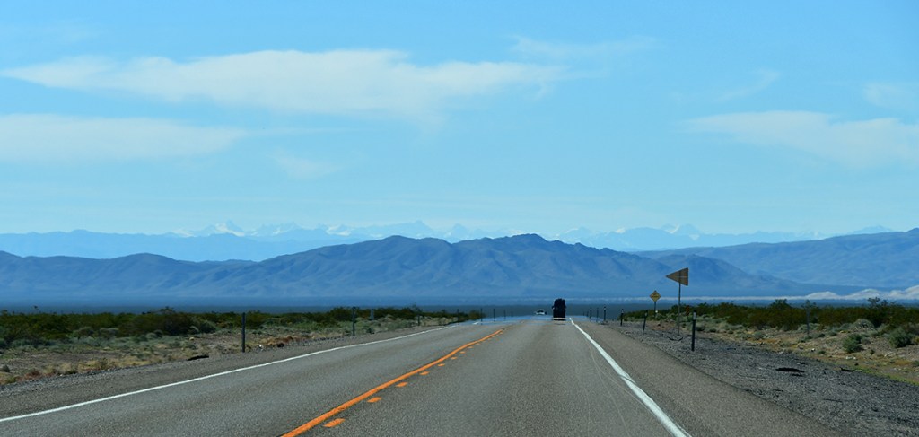 A straight highway stretches into the distance, surrounded by desert terrain and mountains under a clear blue sky.