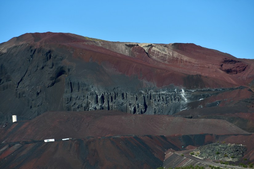 Aerial view of a colorful volcanic landscape featuring layers of red, black, and gray rock against a clear blue sky.