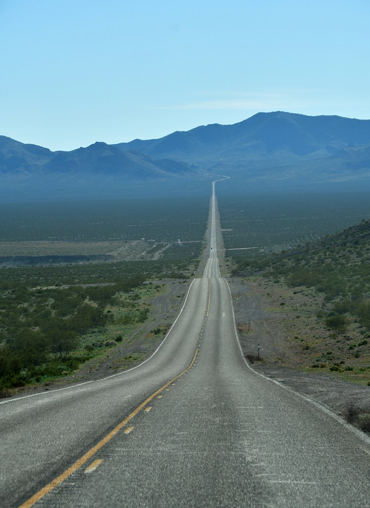 A long, straight road stretches through a desert landscape with mountains in the background under a clear blue sky.