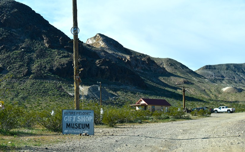 A sign welcoming visitors to a gift shop and museum, located near a dirt road and surrounded by mountainous terrain.