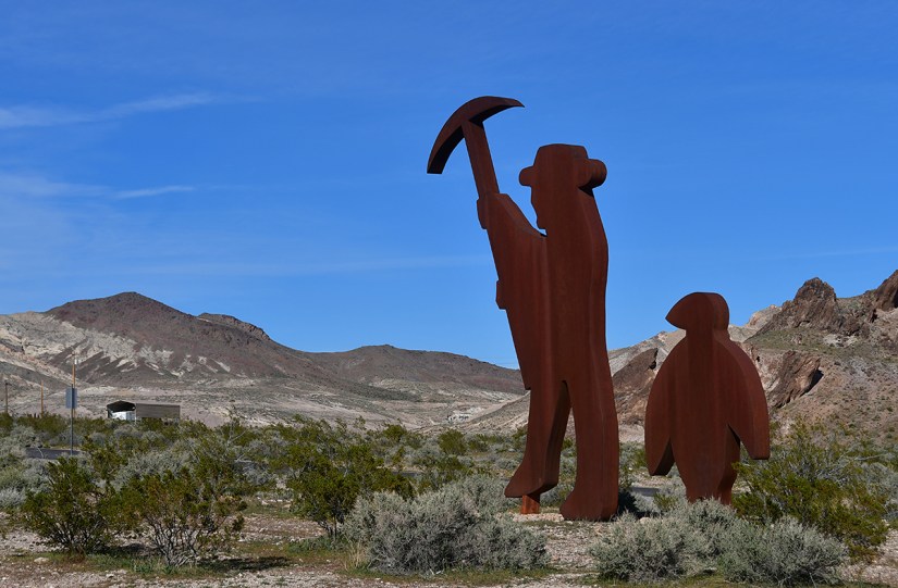 Tall metal sculptures of a prospector with a pickaxe and a smaller figure, set against a desert landscape with mountains in the background.