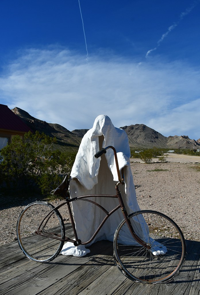 A ghostly figure draped in a white sheet stands next to an old bicycle on a wooden platform with mountains in the background under a blue sky.