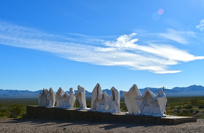 A series of white sculptures depicting robed figures standing on a wooden platform, set against a clear blue sky and distant mountains.