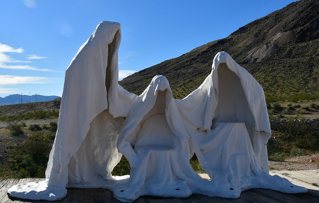 White hooded figures sculpture situated in a desert landscape with mountains in the background.