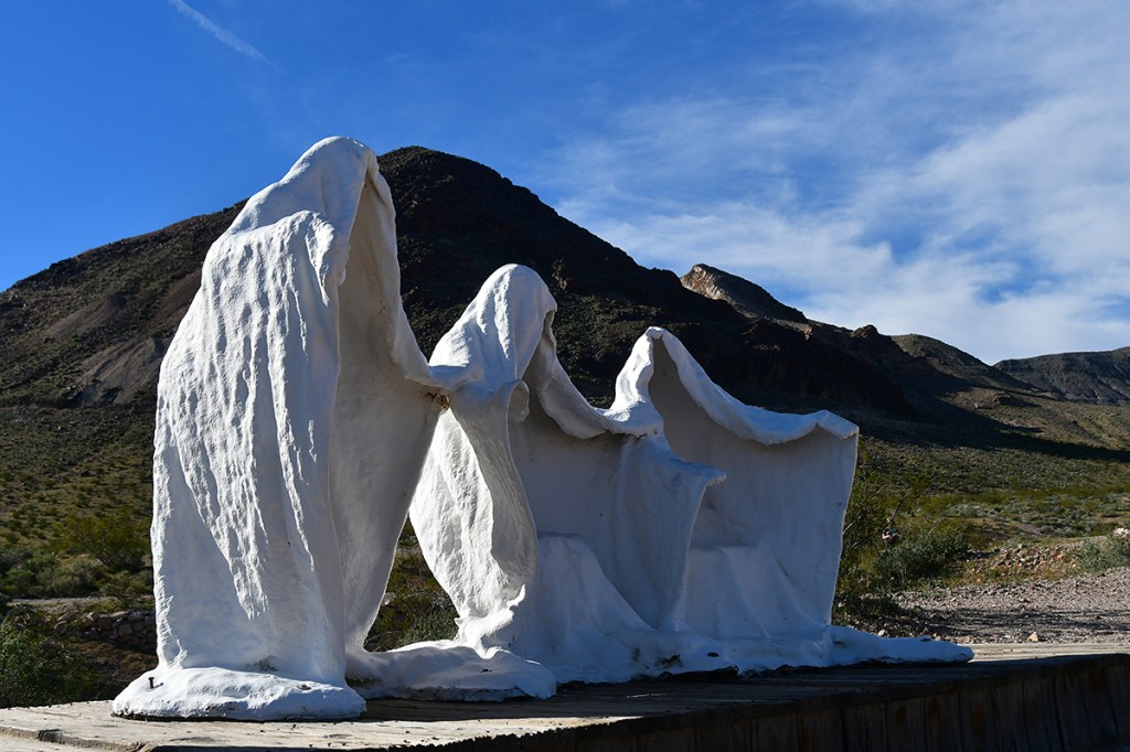Three white, draped sculptures resembling figures, set against a mountainous landscape under a blue sky.