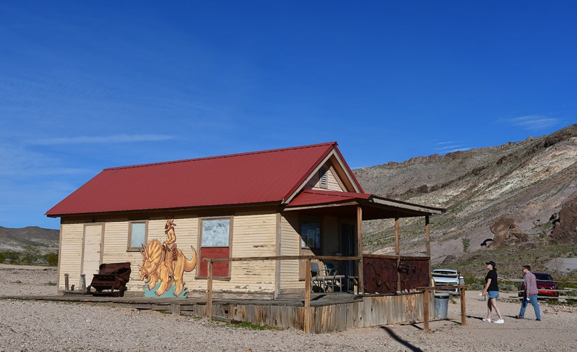 A rustic wooden house with a red metal roof, featuring a colorful mural of a horse on one side. Two people are walking nearby, with mountains in the background and a clear blue sky.
