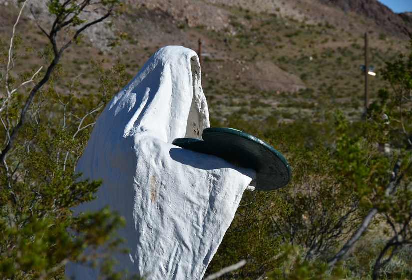 A white statue draped in a ghost-like cloth holding a circular object in a desert landscape with sparse vegetation and mountains in the background.