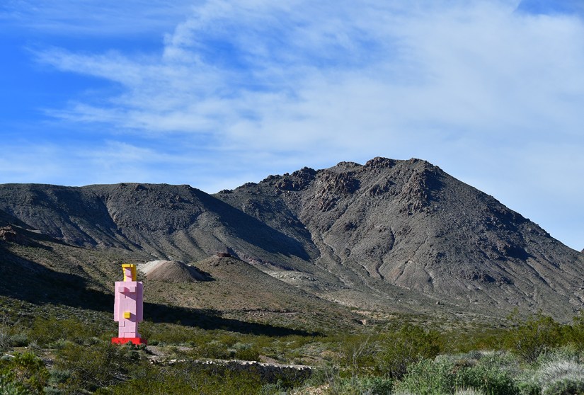 A large pink and yellow sculpture standing in a desert landscape, with mountains in the background and a blue sky.