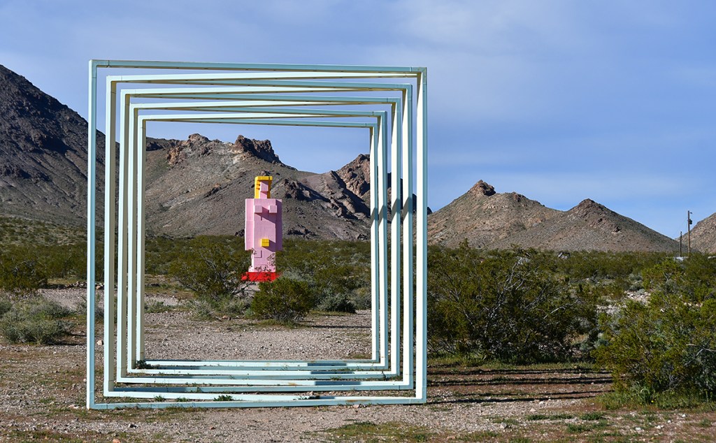 A colorful sculpture of a figure stands in a desert landscape, framed by a series of rectangular structures, with mountains in the background.