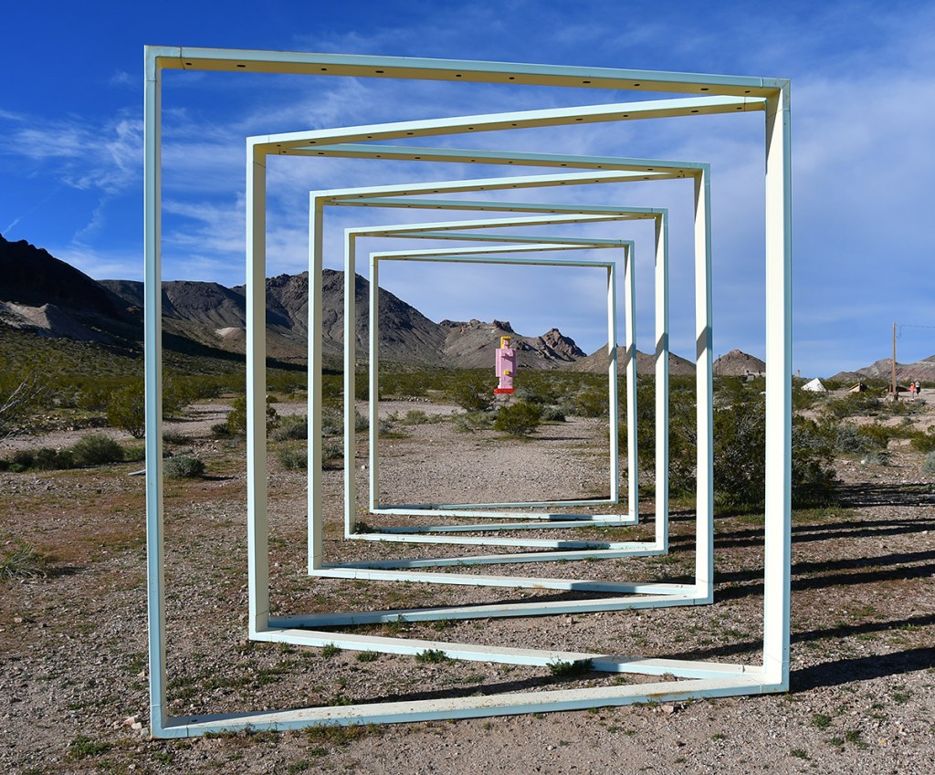A series of white rectangular frames arranged in perspective, leading towards a distant colorful structure in a desert landscape surrounded by mountains.