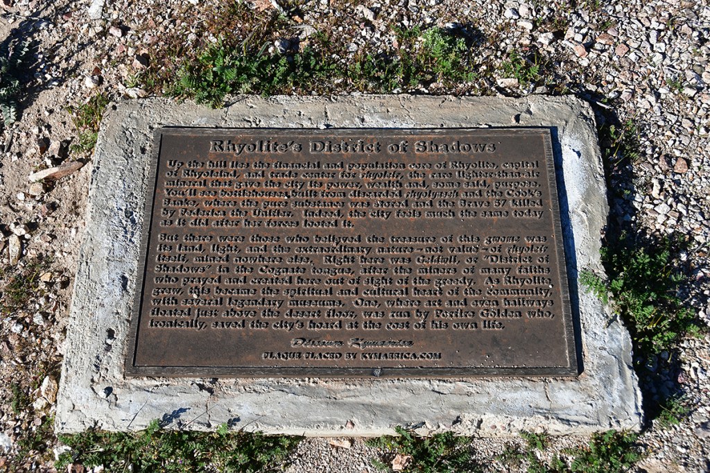 A bronze plaque titled 'Rhyolite's District of Shadows' describing the history and significance of Rhyolite, Nevada, set on a stone base amidst desert vegetation.