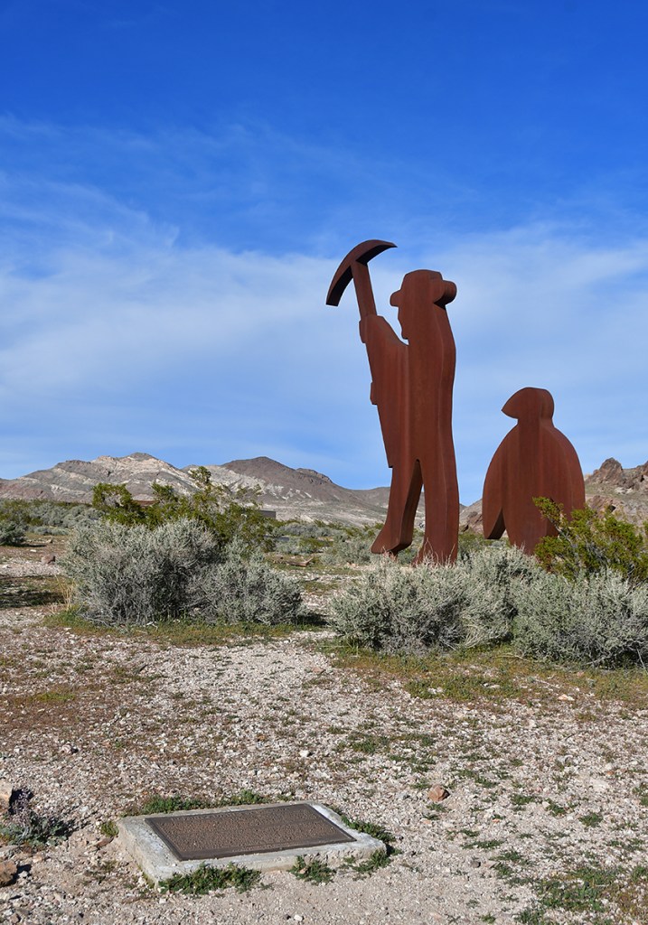 Rustic metal sculptures depicting two figures, one holding a pickaxe, against a backdrop of desert landscape and mountains under a clear blue sky.