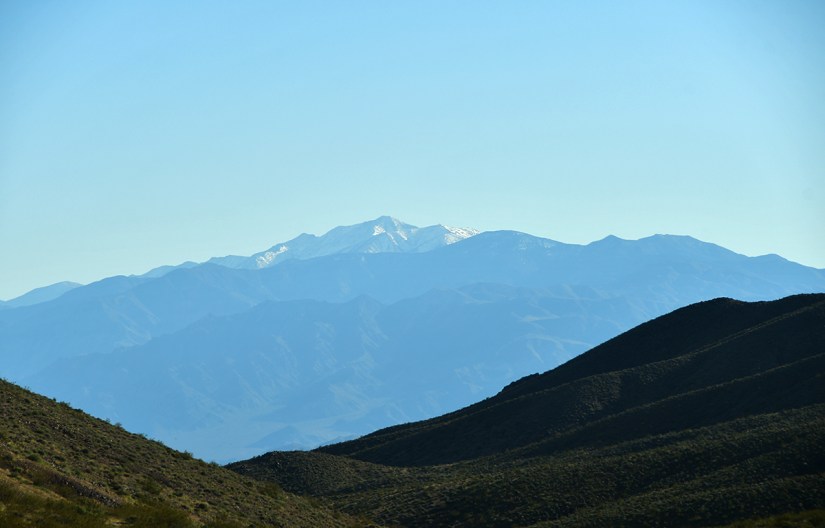 A panoramic view of layered mountains under a clear blue sky, with a snow-capped peak in the background.