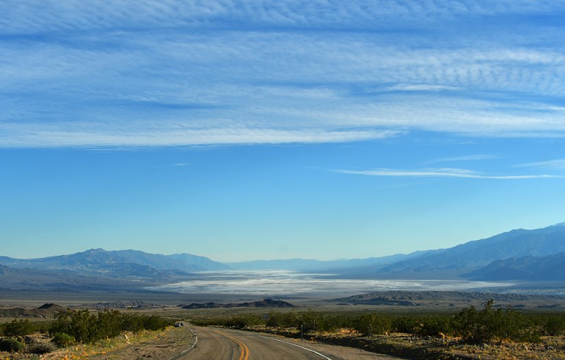 A scenic view of a winding road leading to a vast desert landscape with distant mountains and a blue sky filled with wispy clouds.