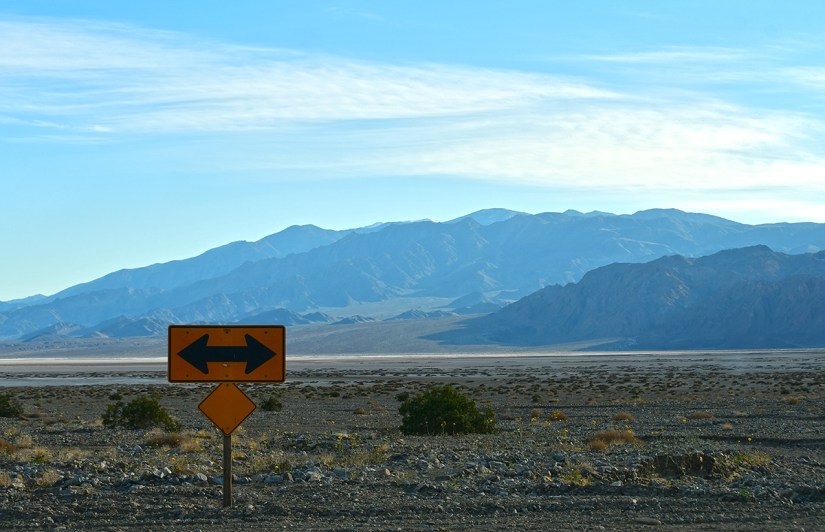 A highway sign indicating a left and right turn with a mountainous landscape in the background under a clear blue sky.