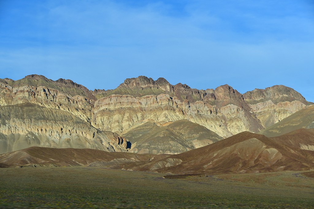 Vast landscape featuring rugged mountains with layered rock formations under a clear blue sky.