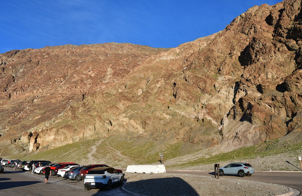 A rocky mountain with colorful layers under a clear blue sky, alongside a parking lot filled with cars.