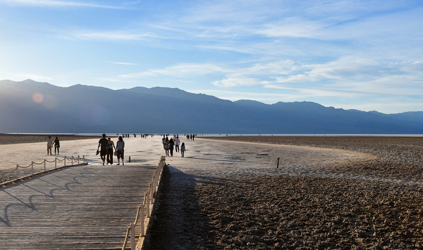 People walking on a wooden boardwalk leading to a vast, dry area with distant mountains under a blue sky.