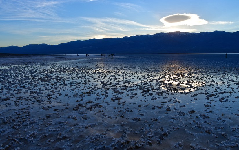 Scenic view of a salt flat at dusk, with reflections on the wet surface, mountains in the background, and people exploring the area.