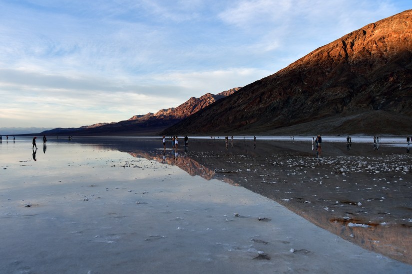 A scenic view of a reflective salt flat during golden hour, with people walking along the surface and mountains in the background.