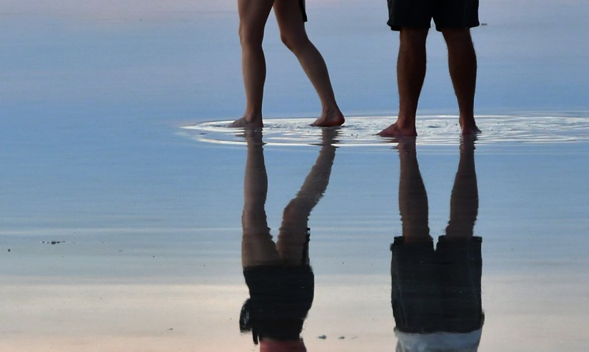 Two people standing barefoot in shallow water, with their reflections visible on the water's surface.