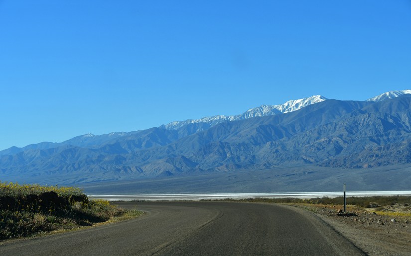 A winding road curves through a mountainous landscape with snow-capped peaks under a clear blue sky.