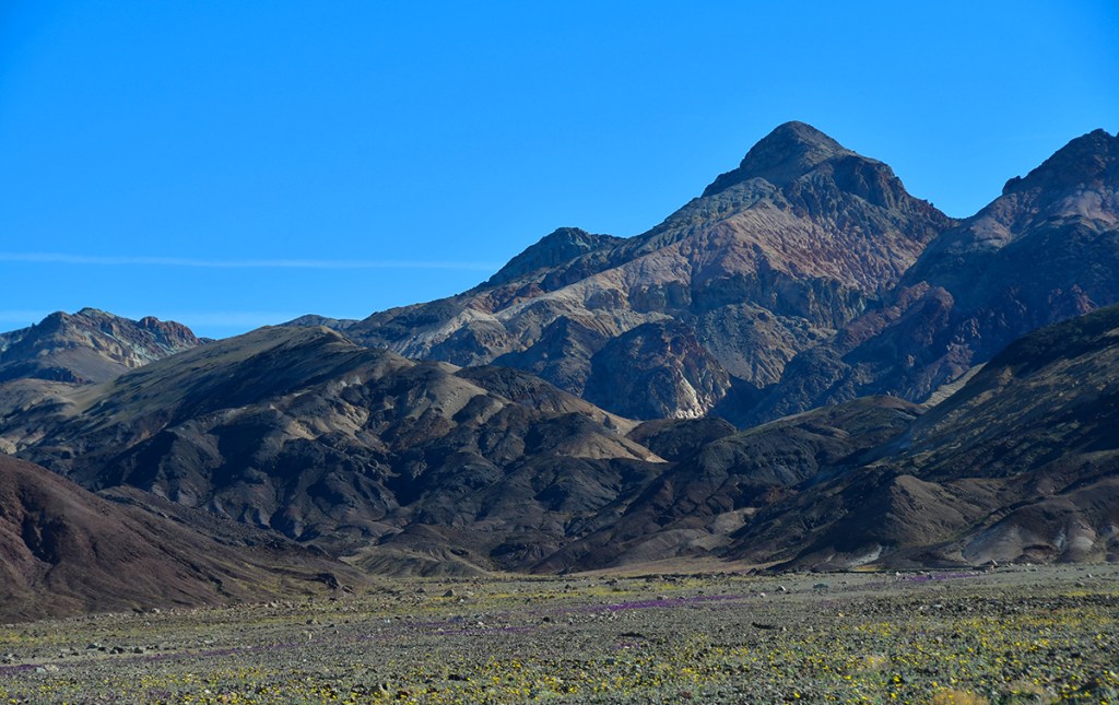 A panoramic view of rugged mountains under a clear blue sky with various shades of earth tones highlighting the rocky terrain.