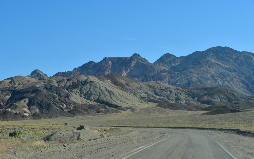 A winding road leads through arid terrain with rugged mountains in the background under a clear blue sky.