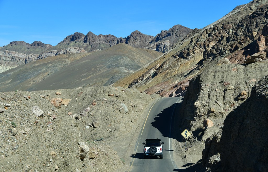 A winding road through a rocky mountain landscape with a white vehicle driving along it under a clear blue sky.