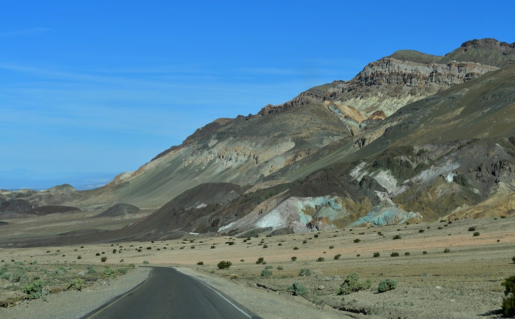 A desert landscape featuring a winding road with colorful rugged mountains in the background and sparse vegetation.