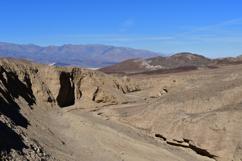 A vast desert landscape featuring dry, rocky terrain, with a clear blue sky and distant mountains in the background.