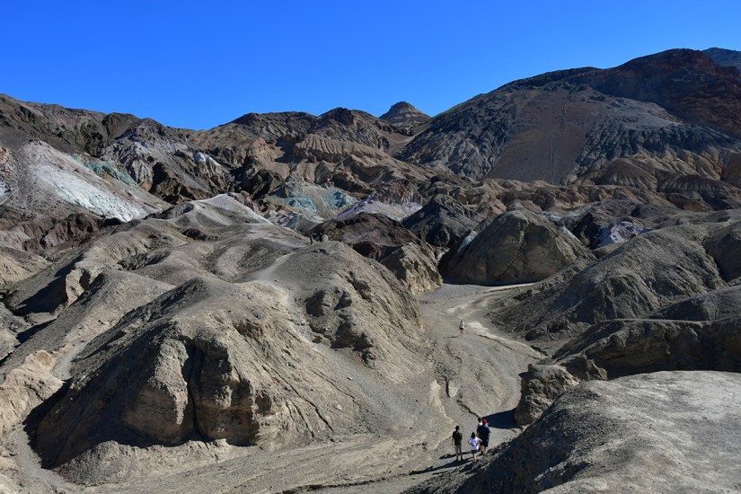Panoramic view of rugged terrain and colorful hills in a desert landscape, with two hikers on a winding path under a clear blue sky.