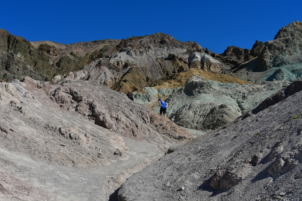 A hiker climbs a rocky terrain with colorful rock formations against a clear blue sky.