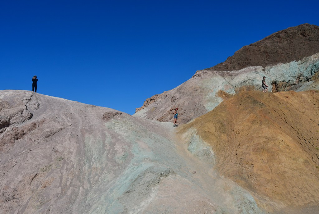 Three people exploring a colorful, textured landscape with rocky formations under a clear blue sky.