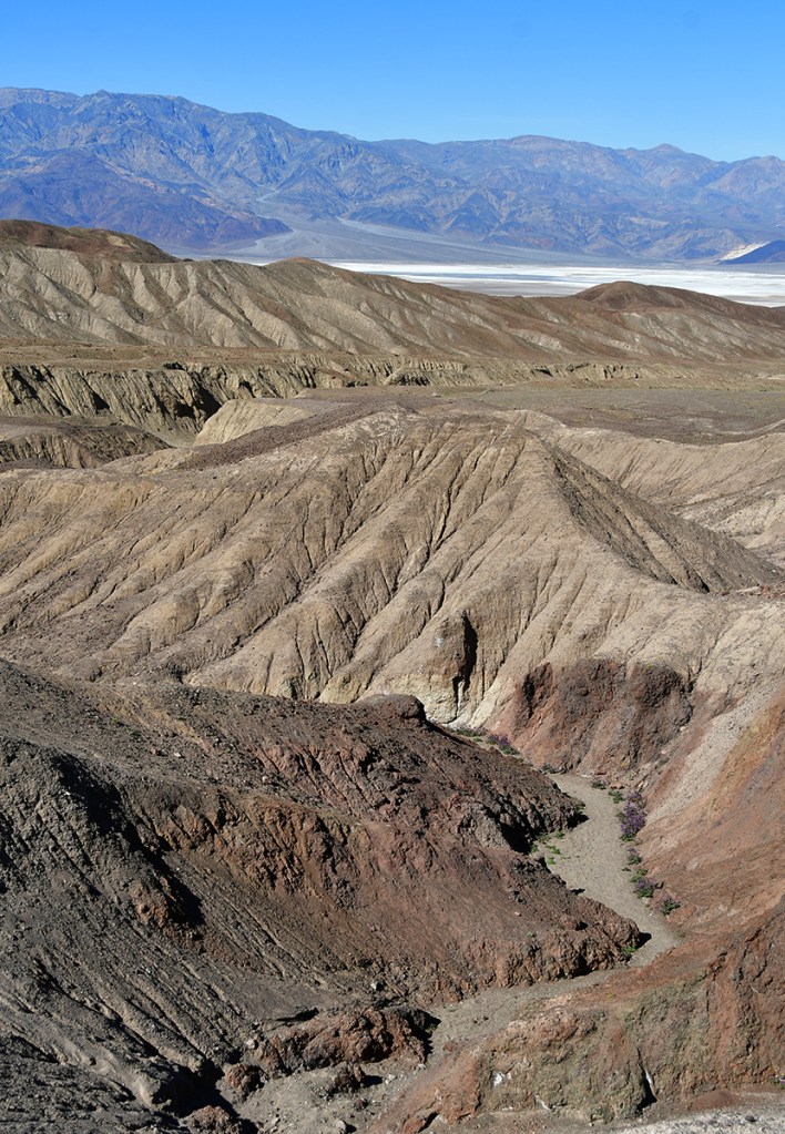 A view of rugged, colorful mountain terrain with undulating ridges and a clear blue sky in the background.