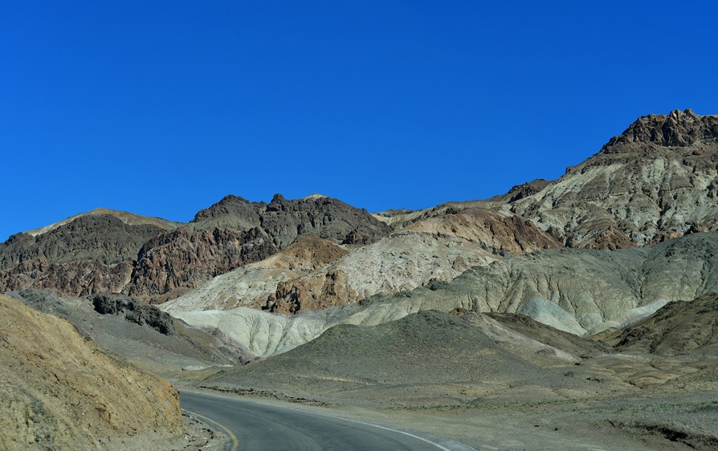 A winding road through arid, multicolored mountains under a clear blue sky.