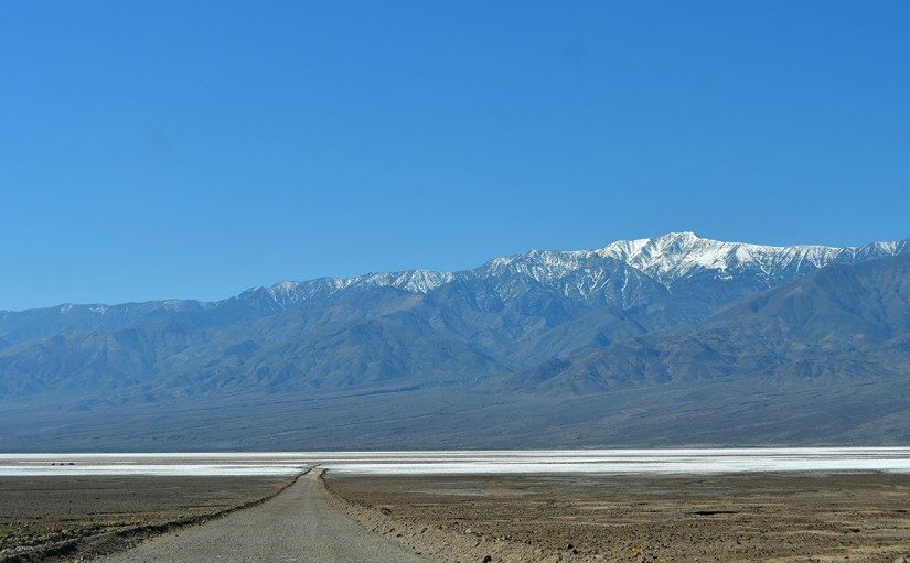 A dirt road leading towards majestic snow-capped mountains under a clear blue sky, with a flat salt pan in the foreground.