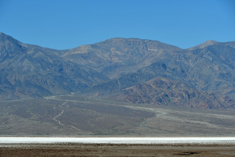 Panoramic view of rugged mountains under a clear blue sky, with a barren landscape in the foreground and a white salt flat visible.