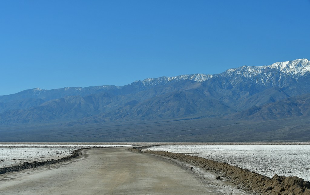 A dirt road winding through a vast salt flat, with distant snow-capped mountains under a clear blue sky.