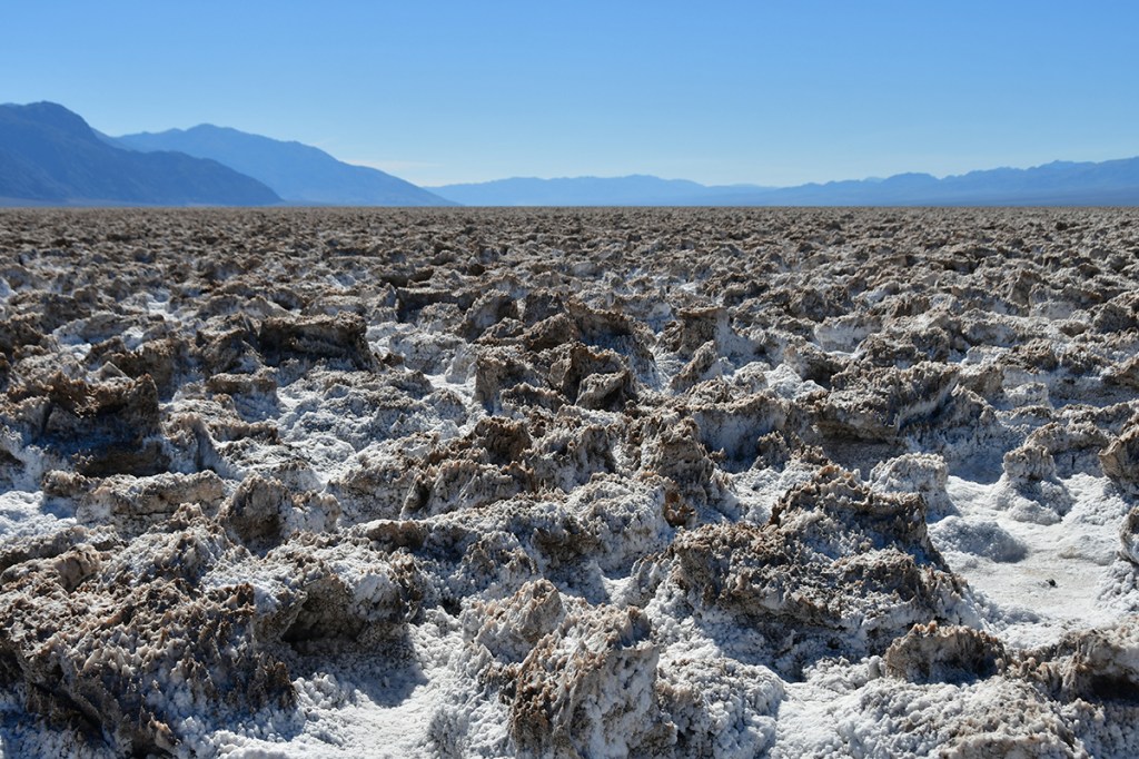A vast salt flat landscape with rugged salt formations under a clear blue sky and distant mountains.