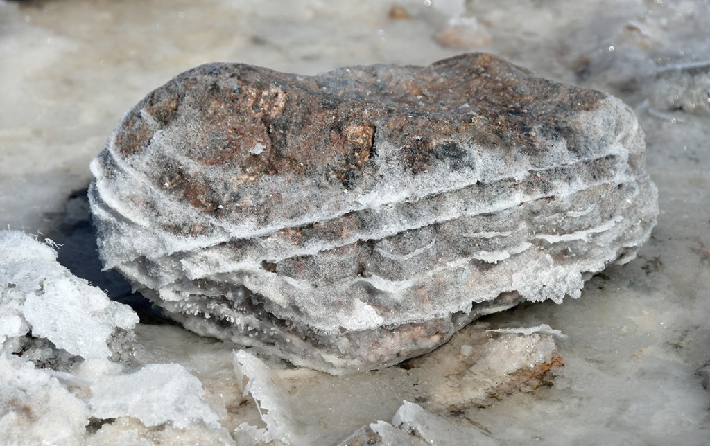 A close-up of a rock with layered formations covered in salt crystals, surrounded by a frosty landscape.
