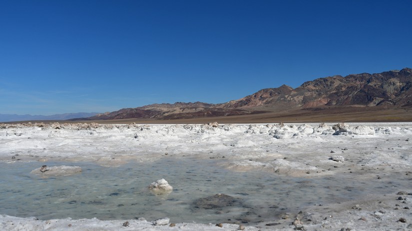 A panoramic view of a salt flat with shallow pools of water, surrounded by rocky mountains under a clear blue sky.