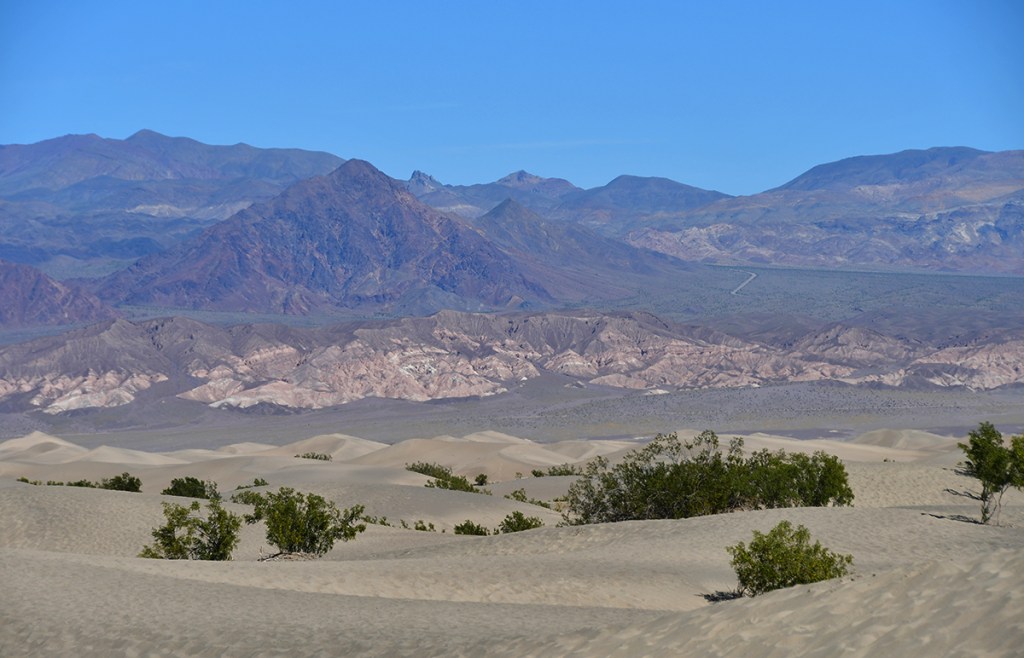A scenic view of desert sand dunes with mountains in the background under a clear blue sky.