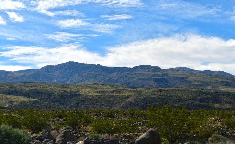 A scenic view of mountains under a blue sky with scattered clouds, featuring green vegetation in the foreground.
