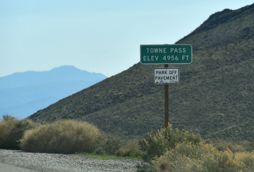 Road sign indicating 'Towne Pass Elev 4956 ft' with an additional sign that says 'Park Off Pavement' next to a hilly landscape.