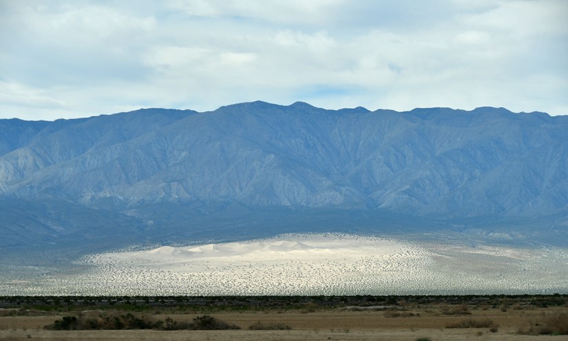 A panoramic view of mountain ranges in the background with a sandy area in the foreground, under a cloudy sky.