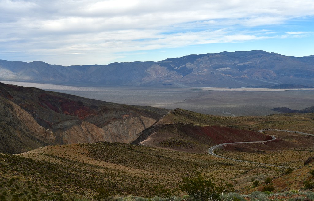 A panoramic view of a desert landscape featuring rugged mountains, colorful rock formations, and a winding road cutting through the terrain under a partly cloudy sky.