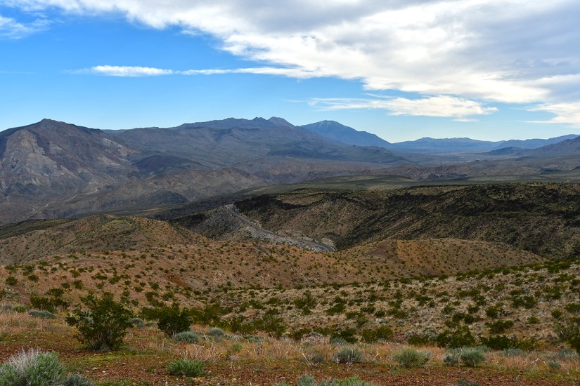 A vast desert landscape featuring rolling hills, rocky mountains, and sparse vegetation under a partly cloudy sky.